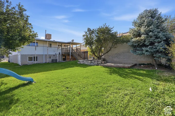 Spacious backyard featuring a manicured lawn, elevated wooden deck, stone-retaining wall, mature landscaping, and an attached staircase