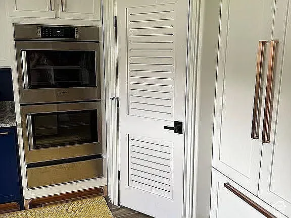 Stainless steel double wall oven, white shaker-style cabinetry, custom pantry door with louvered panels, and metallic finish hardware
