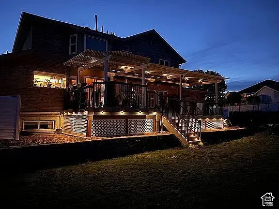 Rear elevation featuring an elevated deck with a pergola, integrated perimeter stair lighting, decorative lattice skirting, and a brick facade