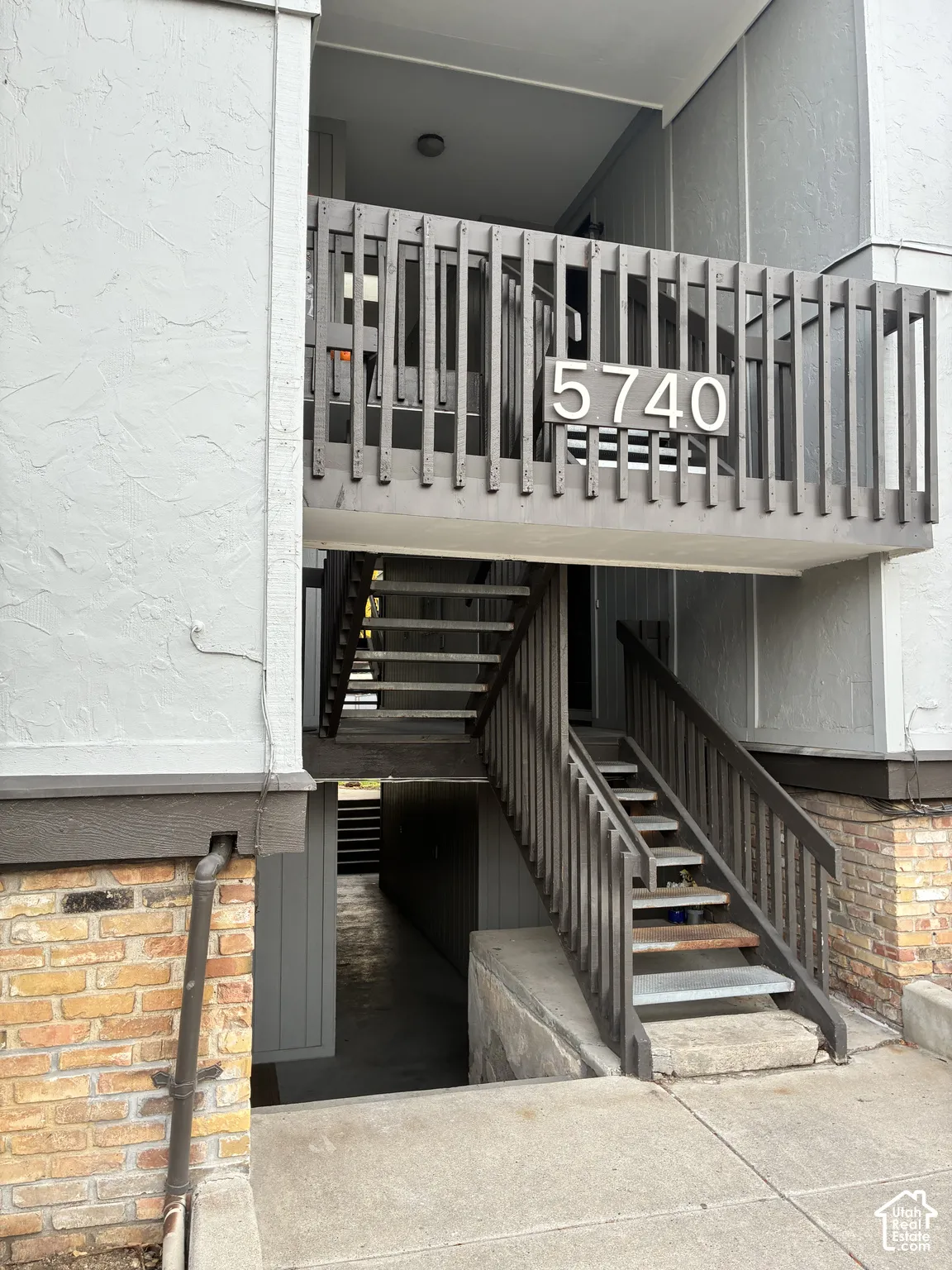Exterior entrance featuring a concrete walkway, multi-level stairs with dark wood-finish railing, textured stucco siding, and decorative brick masonry base