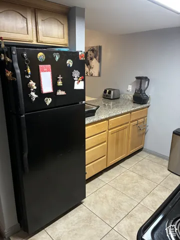Kitchen featuring light-finish wood cabinetry, stone-surface countertops, tile flooring, and a black refrigerator