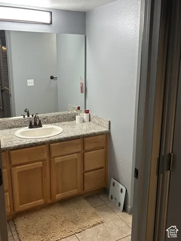 Vanity featuring natural-finish wood cabinetry, stone-composite countertop, oval undermount sink, dual-handle faucet, and full-width wall mirror with integrated lighting fixture