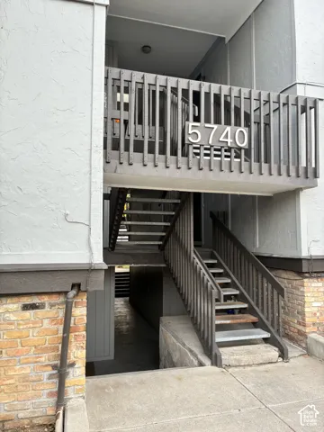 Exterior entrance featuring a concrete walkway, multi-level stairs with dark wood-finish railing, textured stucco siding, and decorative brick masonry base