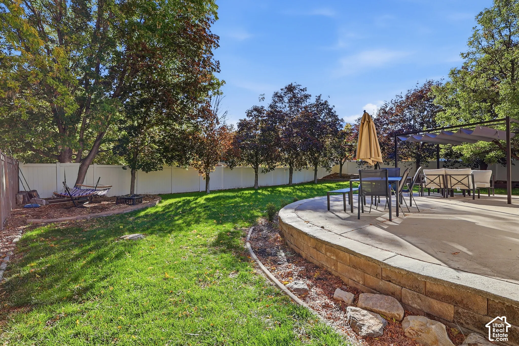Backyard featuring a raised concrete patio, stone-block retaining wall, white vinyl fencing, and mature trees