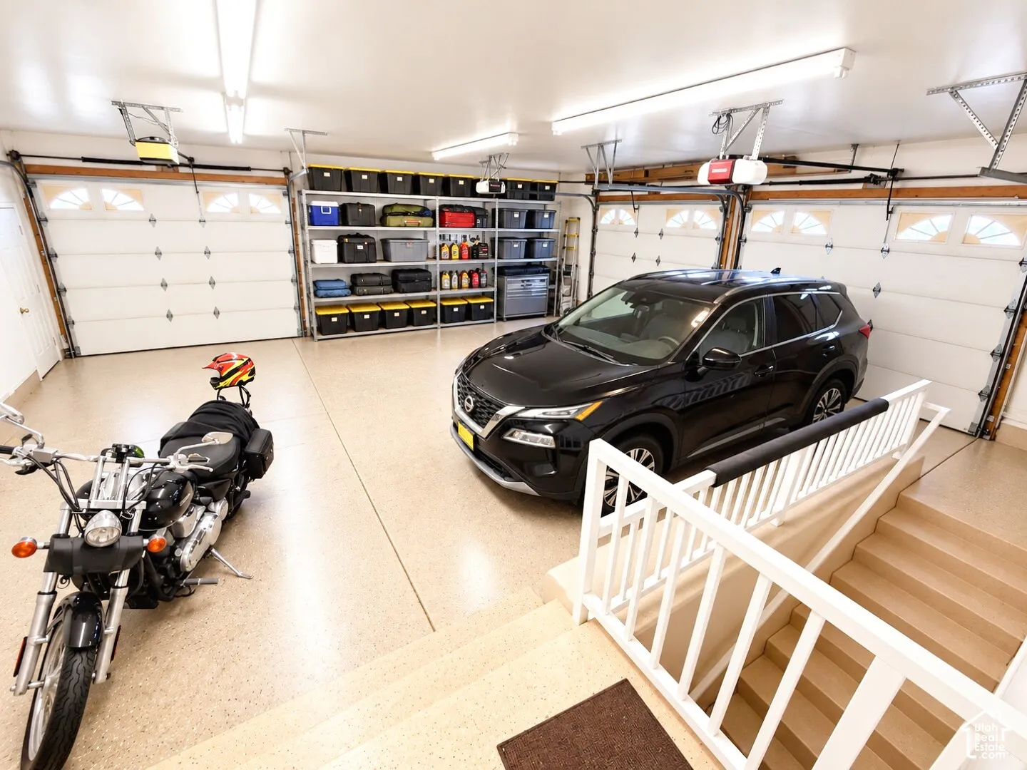 Attached two-car garage featuring epoxy-coated flooring, dual automatic garage door openers with inset windows, built-in metal shelving, and white railings surrounding the stairwell