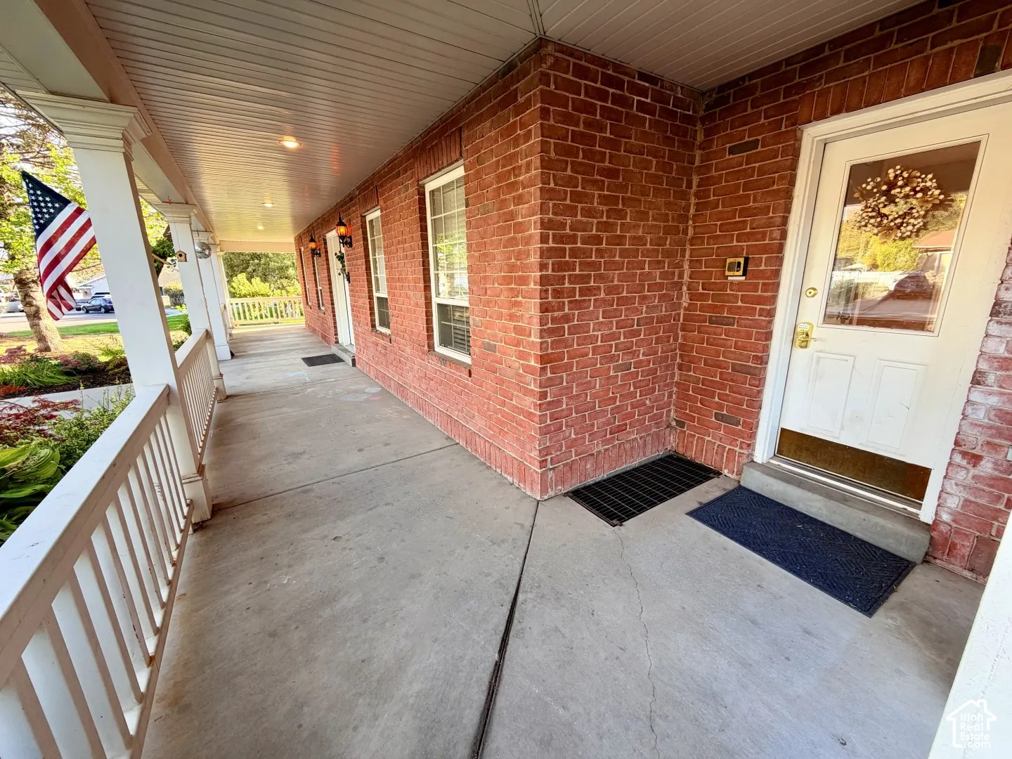 Covered front porch featuring brick siding, a concrete floor, white spindle railings, and wall-mounted exterior lighting