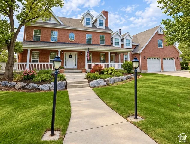 Traditional red brick exterior featuring a covered wraparound porch, gabled roofline with multiple dormers, two-car garage, and stone landscape curbing