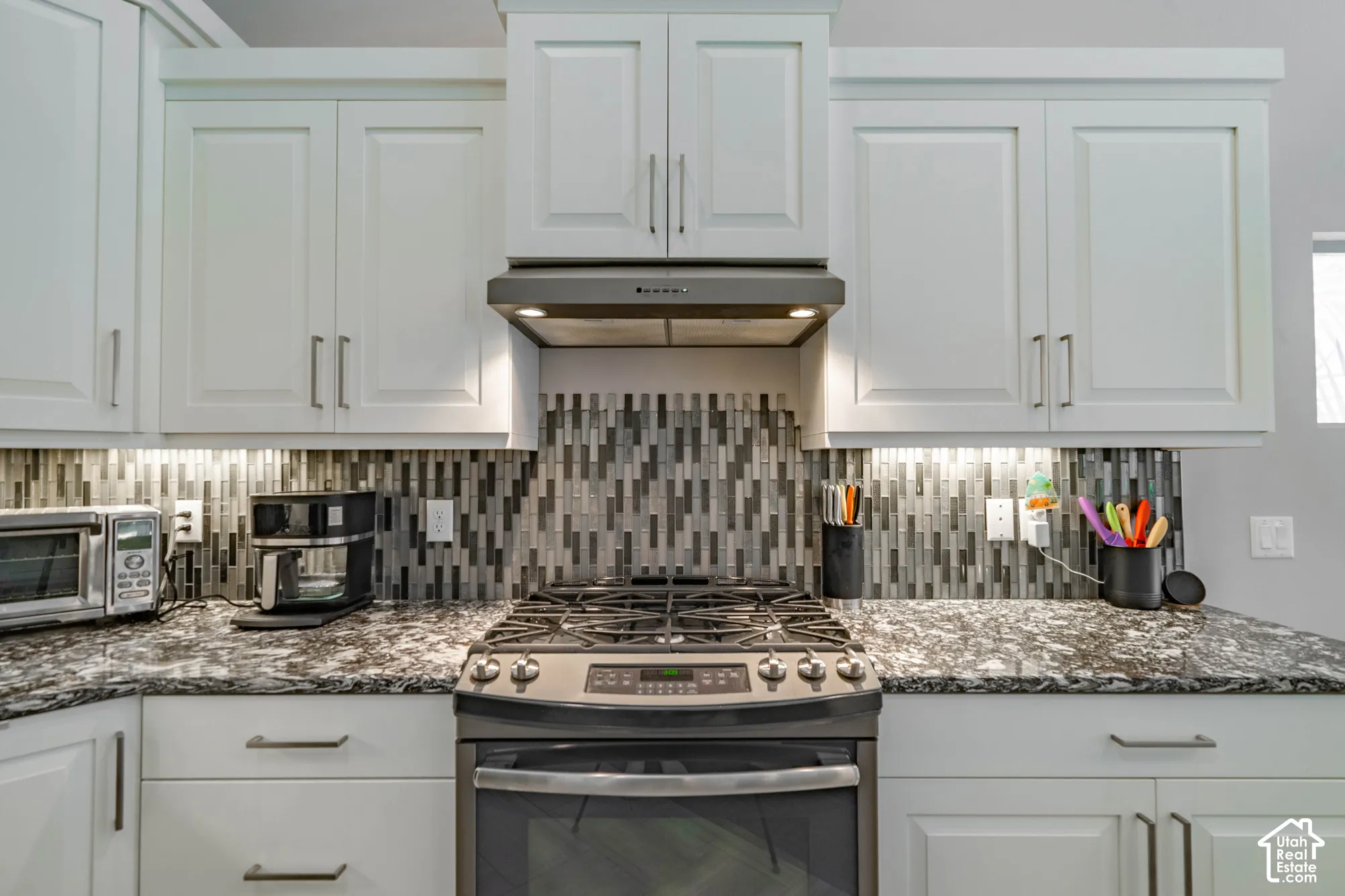 White shaker-style cabinetry with satin nickel pulls, stone countertop, multi-tonal linear tile backsplash, and stainless steel gas range with a matching range hood