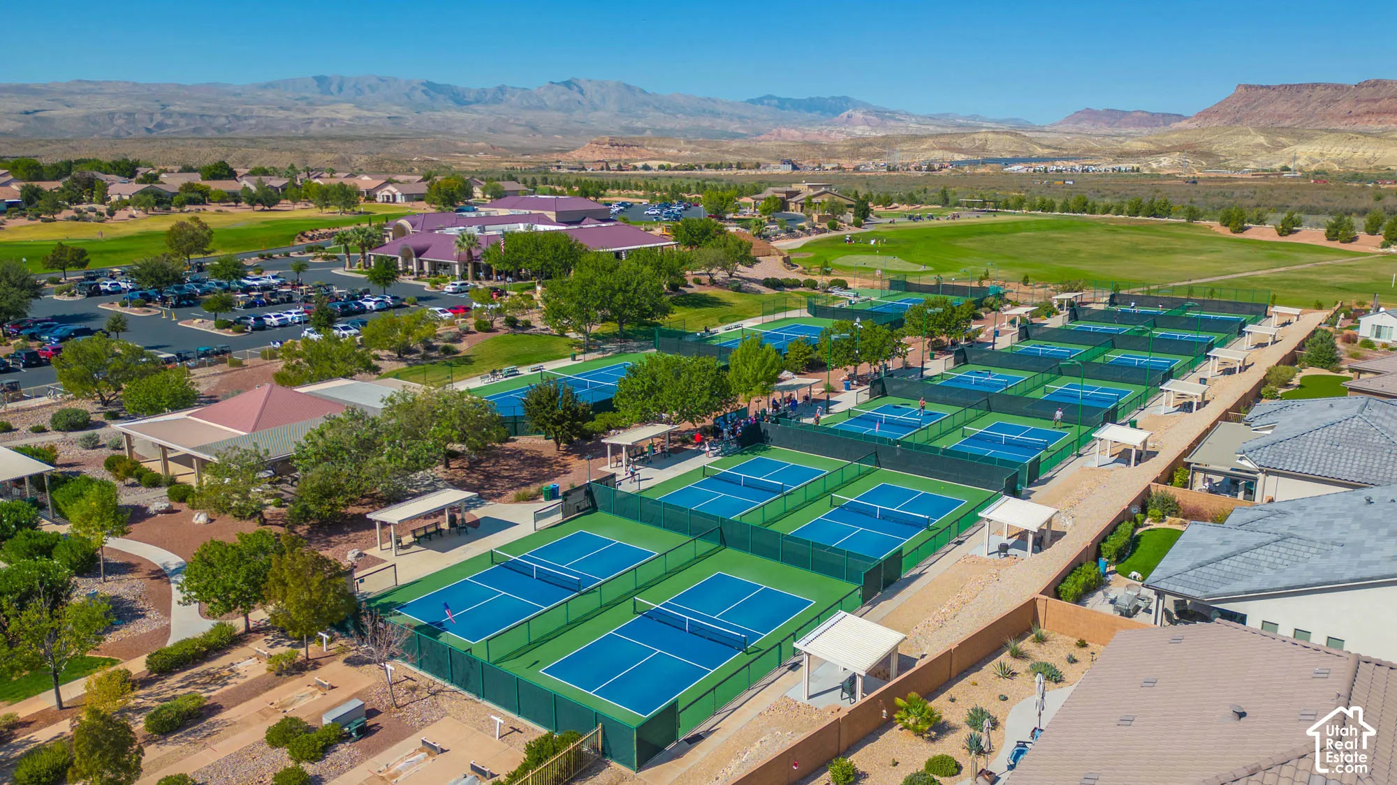 Aerial perspective of a recreation complex featuring multiple fenced tennis and pickleball courts, shaded pavilions, paved pedestrian walkways, and desert landscape with mountain scenery