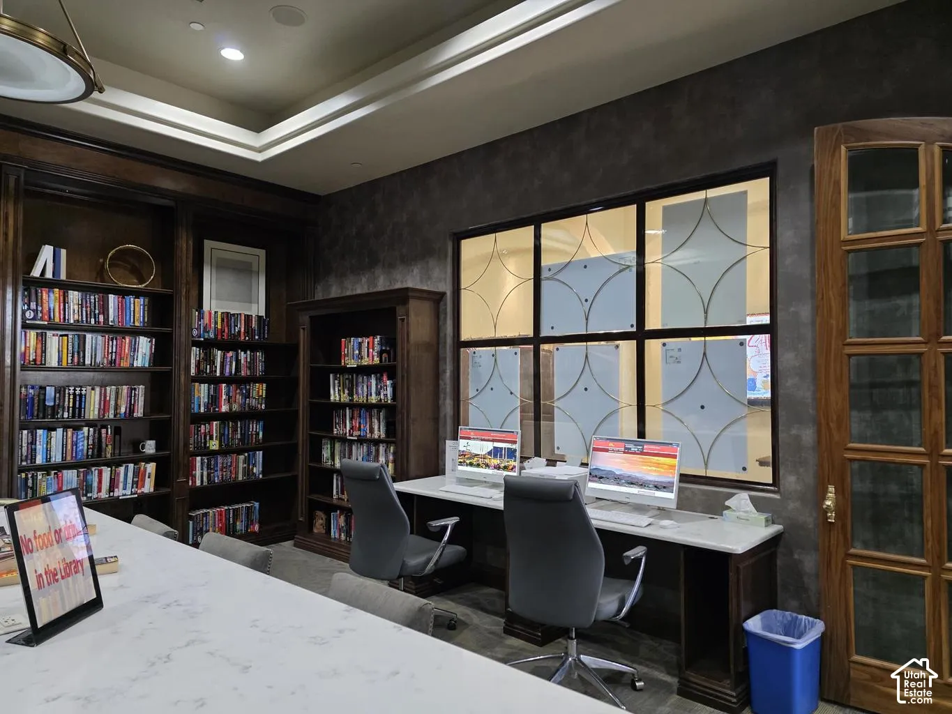 Custom floor-to-ceiling wooden bookshelves, tray ceiling with recessed lighting, stone-topped work surfaces, interior window with geometric frosted detailing, and multi-pane wood door