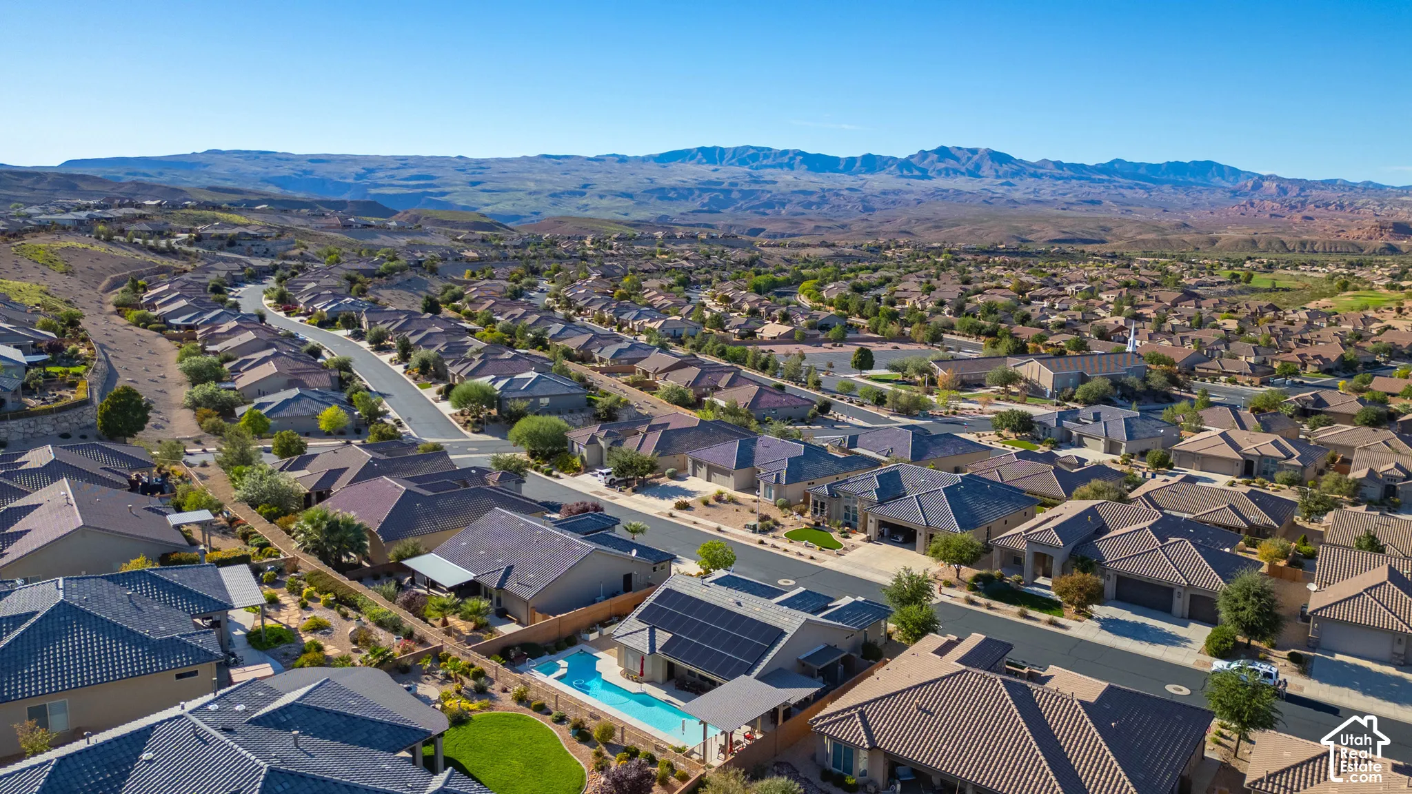 Aerial perspective of a residential property featuring an in-ground swimming pool, solar panel array, covered patio, and manicured landscaping, set against a desert mountain backdrop