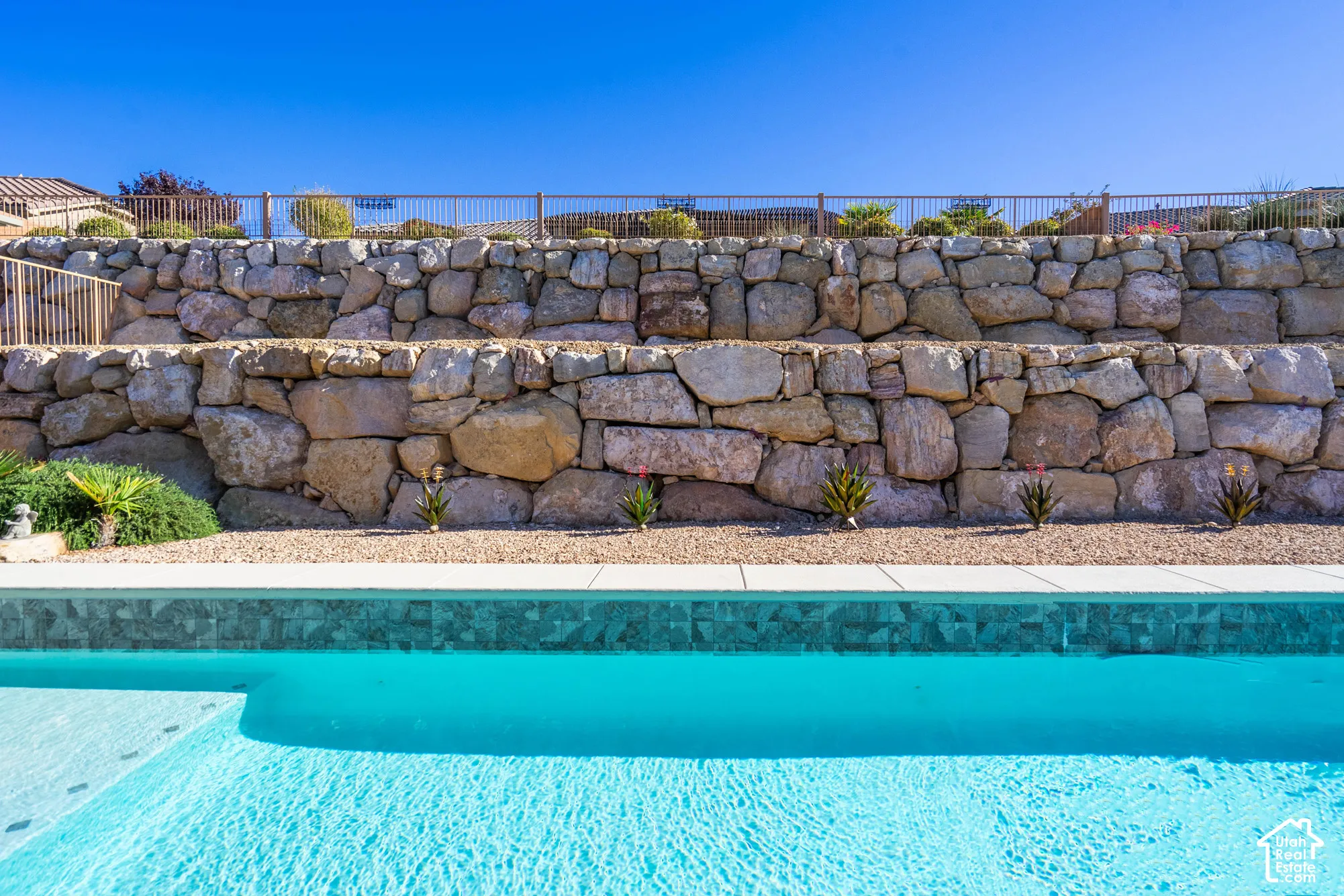 Swimming pool featuring tiled coping and light-toned surfacing, backed by a multi-tiered natural stone retaining wall and metal perimeter fencing