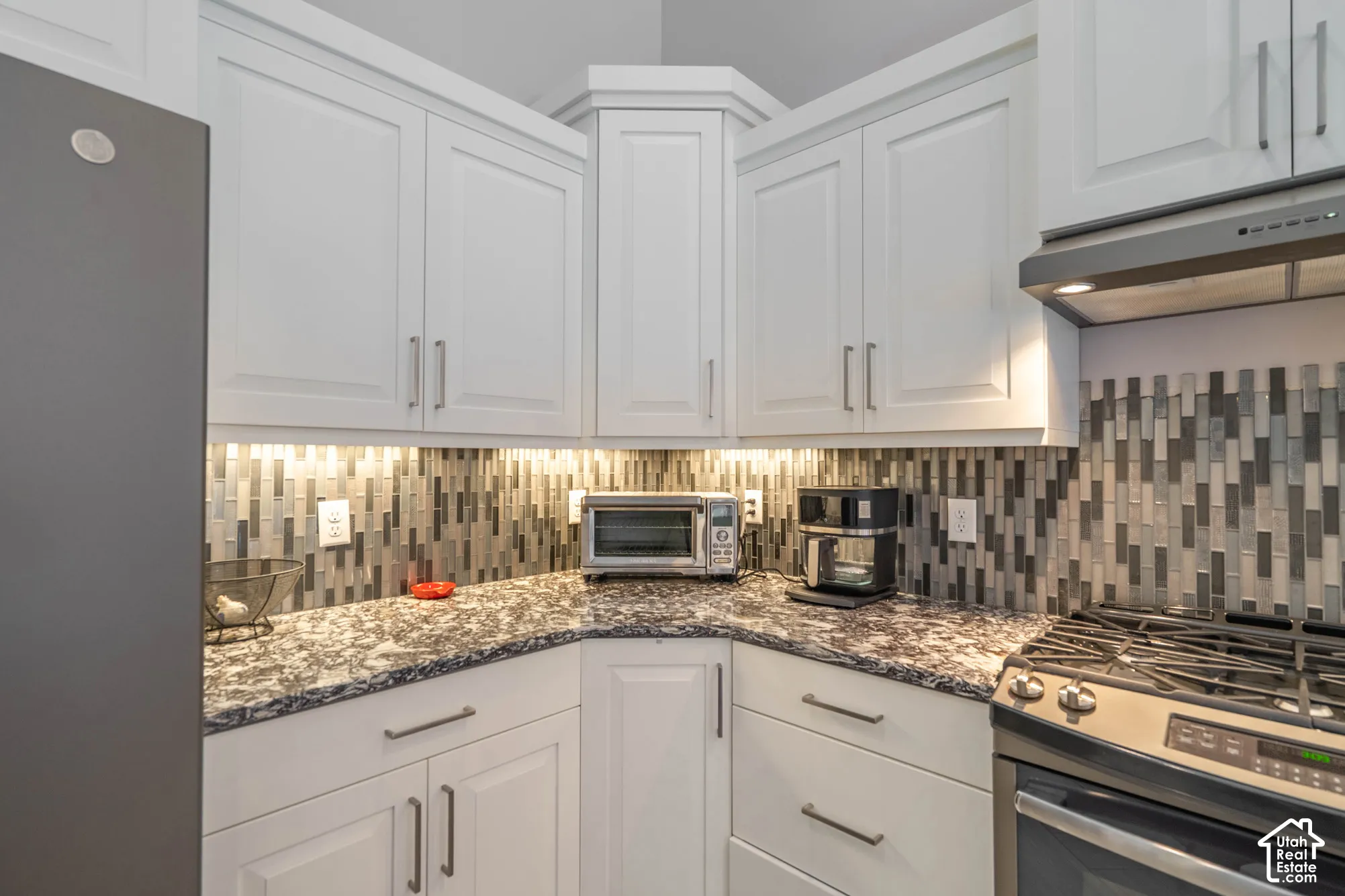 White shaker-style cabinetry with long metallic pulls, patterned stone-finish countertops, multicolored vertical tile backsplash, under-cabinet lighting, and stainless steel gas range with integrated vent hood