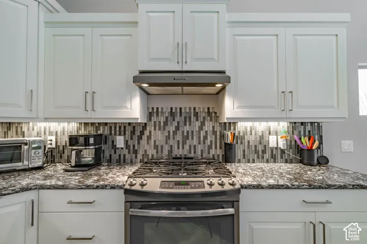 White shaker-style cabinetry with satin nickel pulls, stone countertop, multi-tonal linear tile backsplash, and stainless steel gas range with a matching range hood