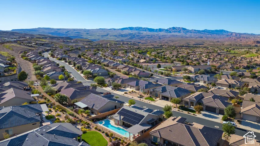 Aerial perspective of a residential property featuring an in-ground swimming pool, solar panel array, covered patio, and manicured landscaping, set against a desert mountain backdrop