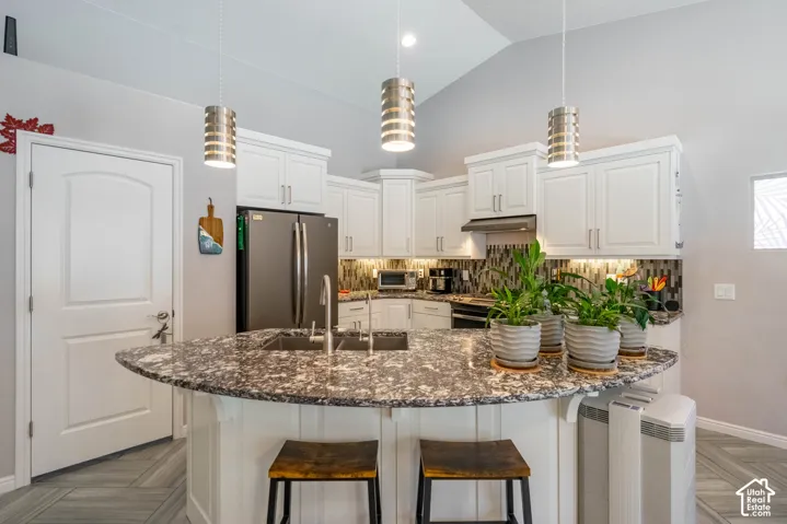 Kitchen featuring white cabinetry, stone-surfaced center island with a built-in sink, multi-toned tile backsplash, and three metallic pendant light fixtures