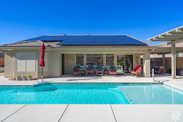 In-ground swimming pool featuring a Baja shelf, concrete decking, roof-mounted solar panels, and a covered patio structure