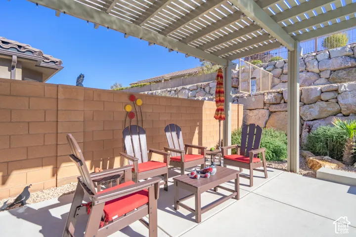 Covered concrete patio featuring a louvered overhead structure, tan masonry block perimeter wall, and tiered natural stone retaining wall