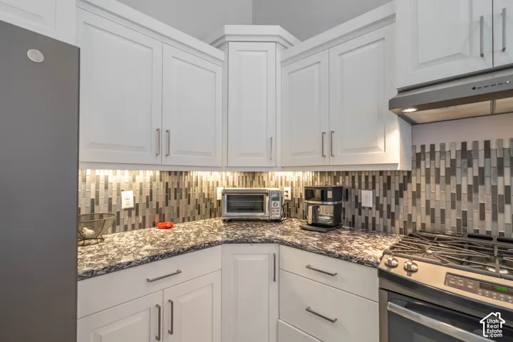 White shaker-style cabinetry with long metallic pulls, patterned stone-finish countertops, multicolored vertical tile backsplash, under-cabinet lighting, and stainless steel gas range with integrated vent hood