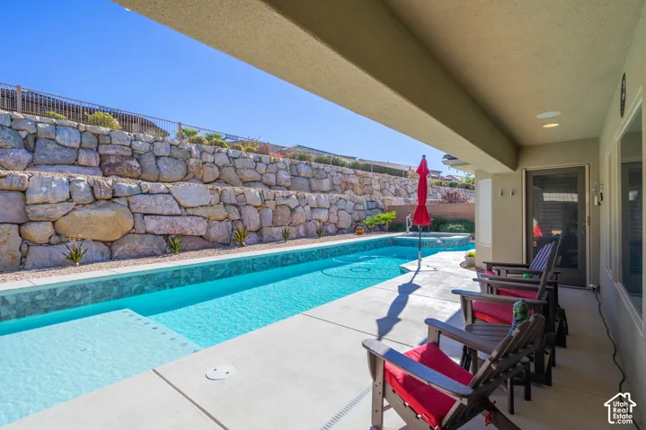 Covered concrete patio featuring a rectangular swimming pool with integrated spa and decorative tiered stone retaining wall