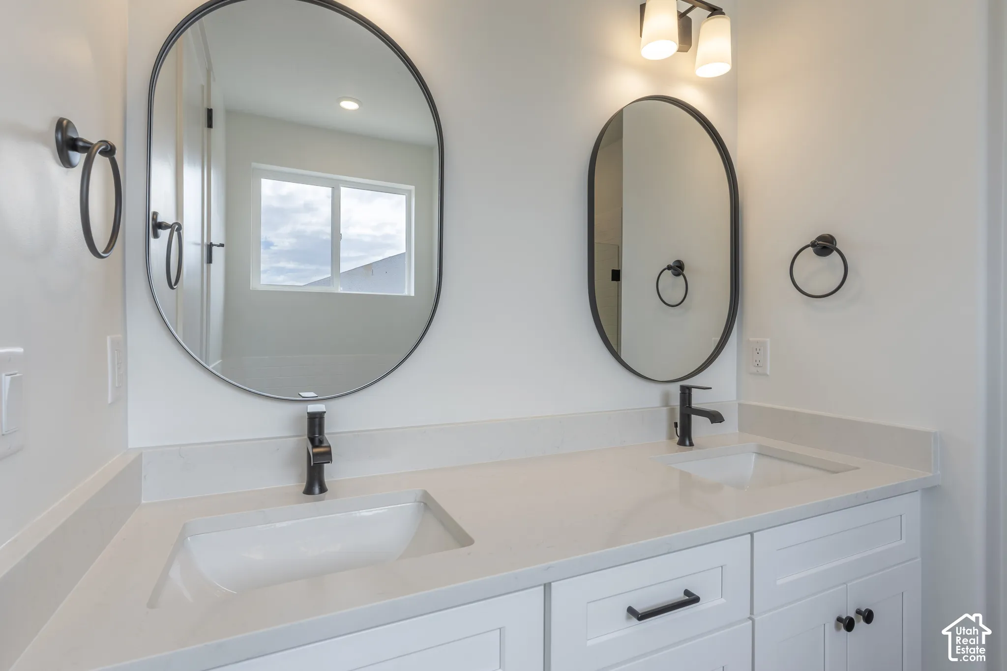 Double vanity featuring white cabinetry, solid surface countertop with integrated dual sinks, matte black hardware, oval mirrors, and wall-mounted light fixtures