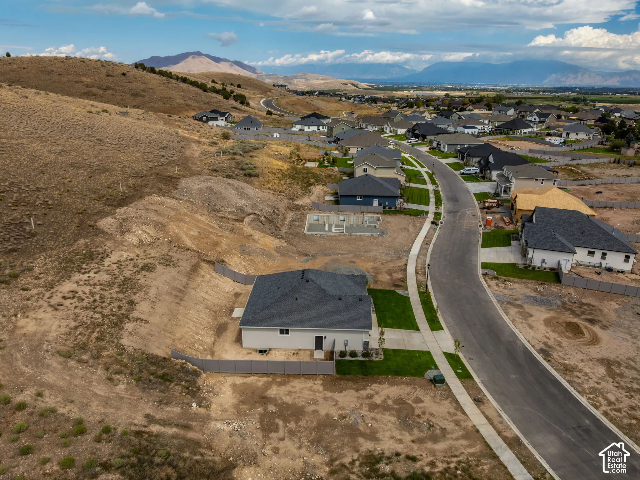 Aerial perspective of a residential property featuring a dark-shingled roof, attached concrete driveway, perimeter fencing, and expansive mountain scenery