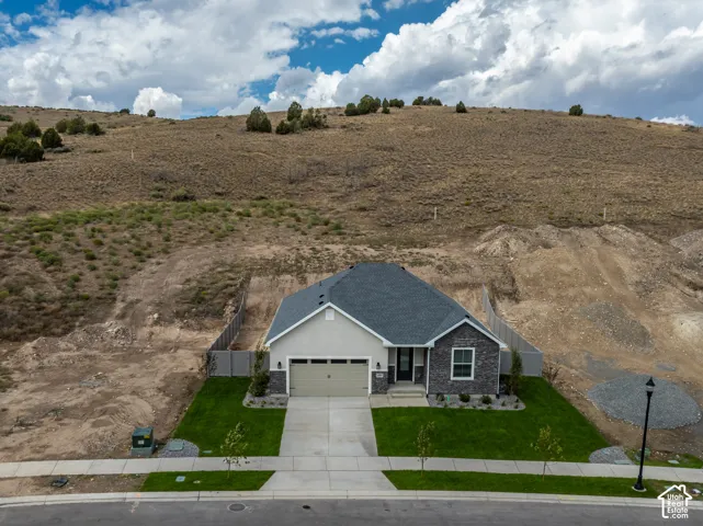 Single-story residence featuring a two-car garage, stone veneer siding, concrete driveway, and gray shingled roof, set against an expansive hillside backdrop