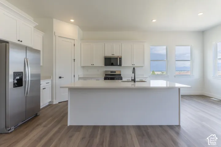 Kitchen featuring white shaker-style cabinetry, quartz countertops, a central island with an integrated sink, stainless steel appliances, and wood-finish flooring