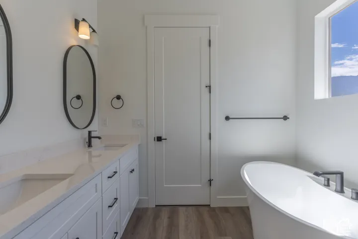 Double vanity with a light-toned solid surface countertop, oval mirrors, matte black hardware, freestanding soaking tub, and wood-finish flooring