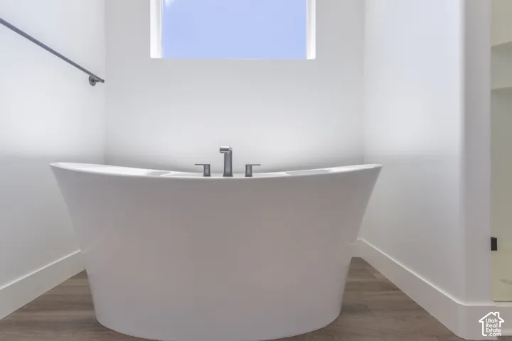Freestanding white soaking tub with a deck-mounted metal faucet, wood-finish flooring, and a window opening
