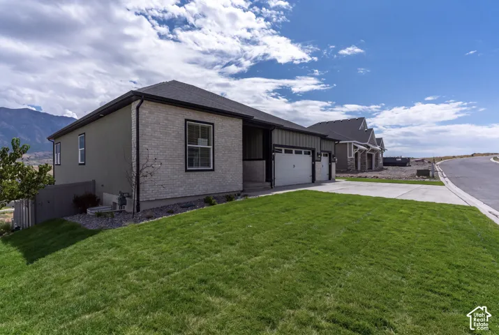 Single-story exterior featuring a light-colored brick veneer, dark vertical siding, concrete driveway, attached multi-car garage, and mountain backdrop