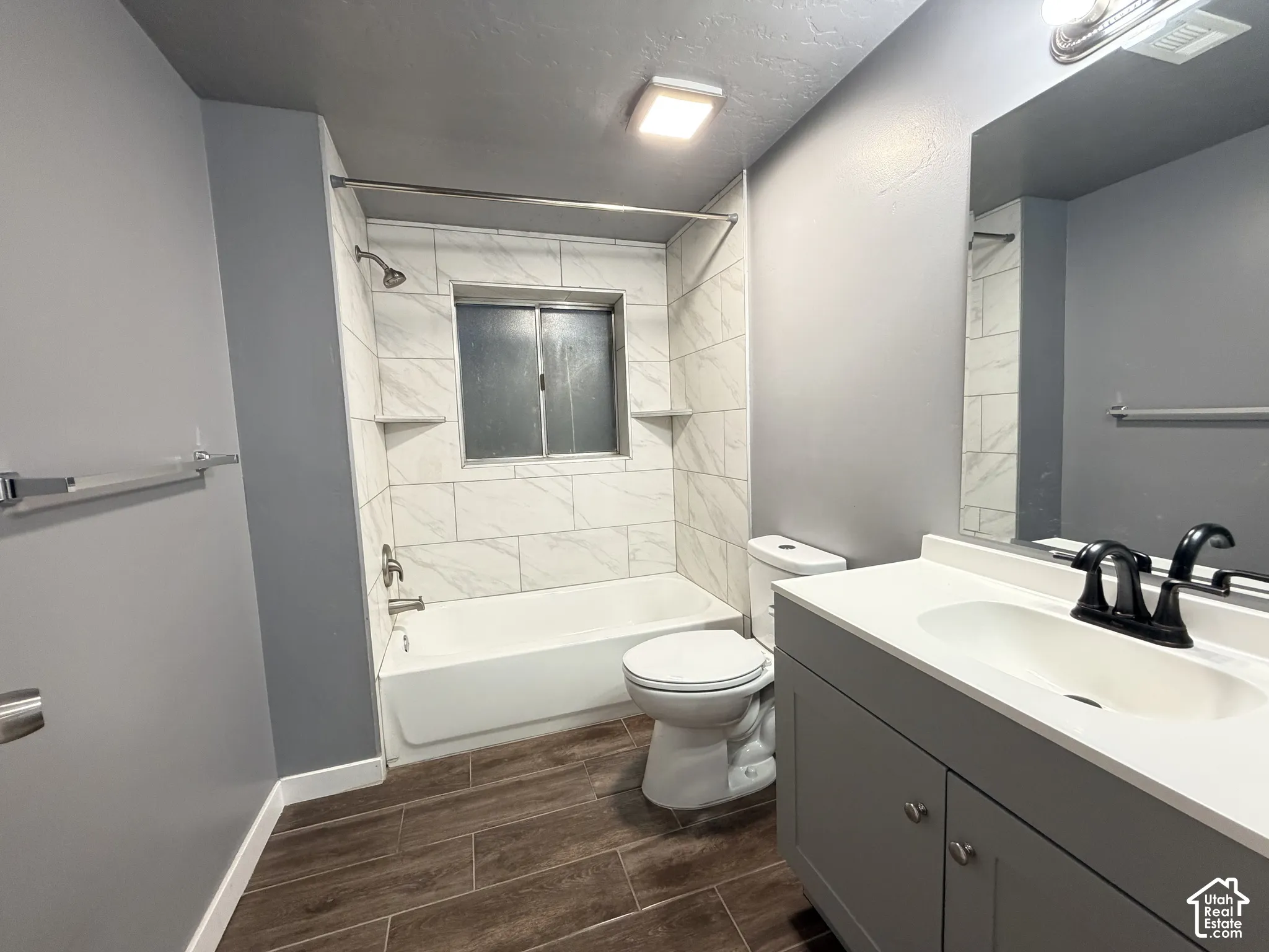 Full bathroom featuring wood-finish flooring, a vanity with a white countertop and matte black dual-handle faucet, a large wall-mounted mirror, and a tub-shower combination with floor-to-ceiling tile surround