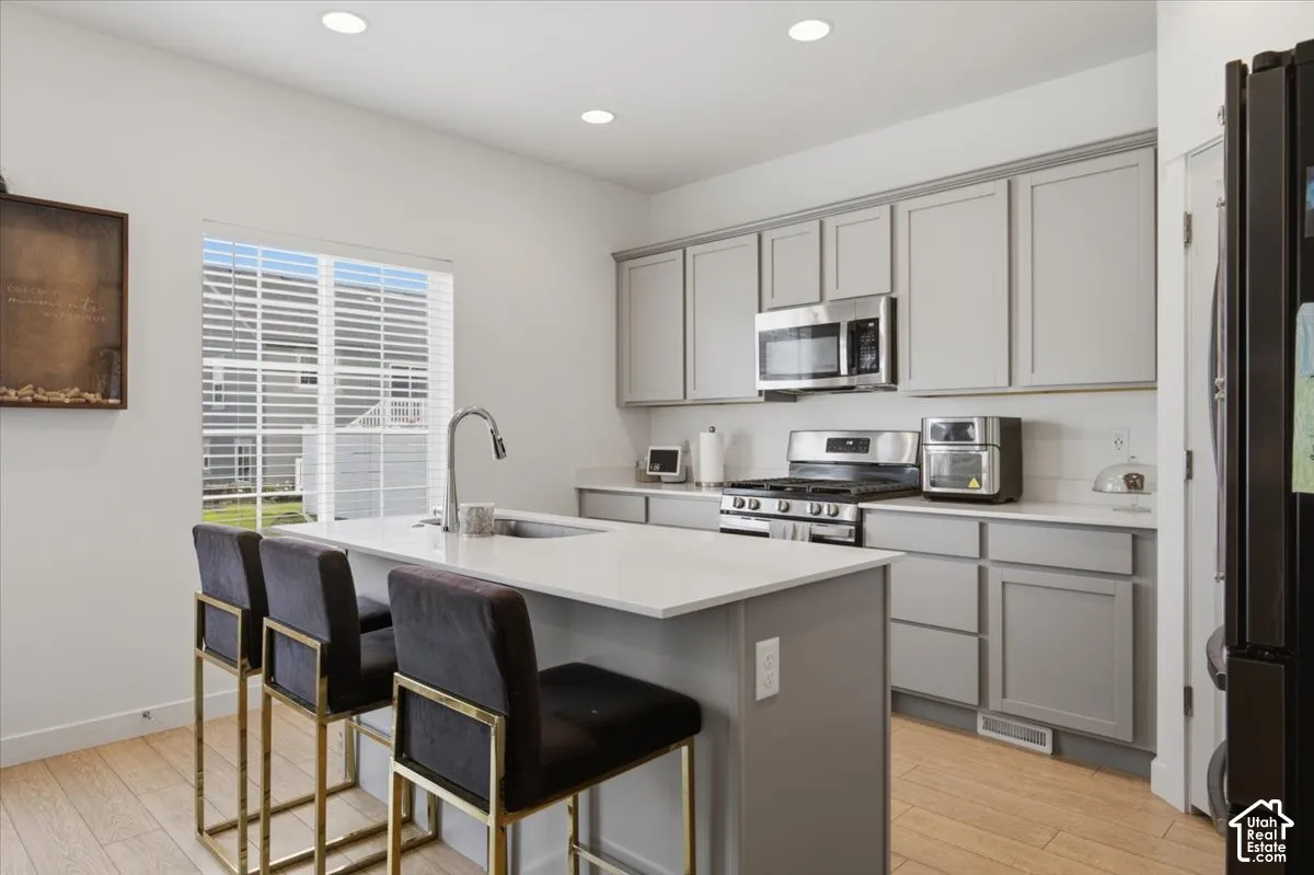 Kitchen featuring grey shaker-style cabinetry, quartz countertops, a central island with a stainless steel sink, stainless steel appliances, and wood-finish flooring