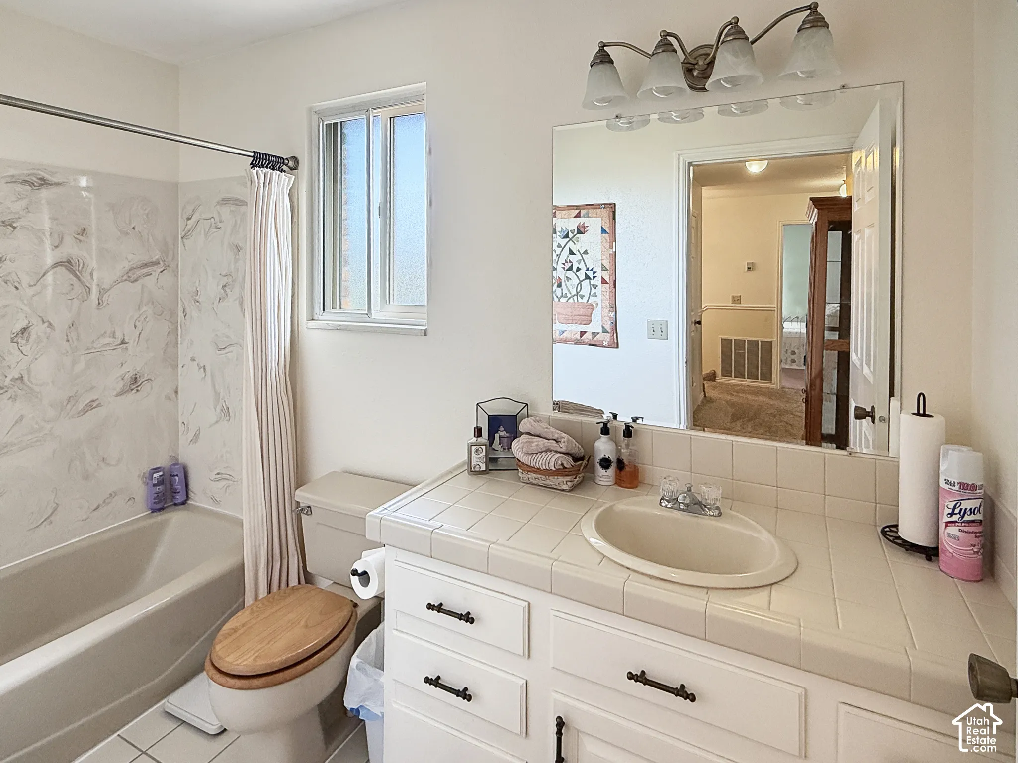 Bathroom featuring a tiled vanity countertop with an integrated oval sink, matte-finish cabinetry, large wall-mounted mirror, four-light vanity fixture, and a tub-shower combination with decorative surround
