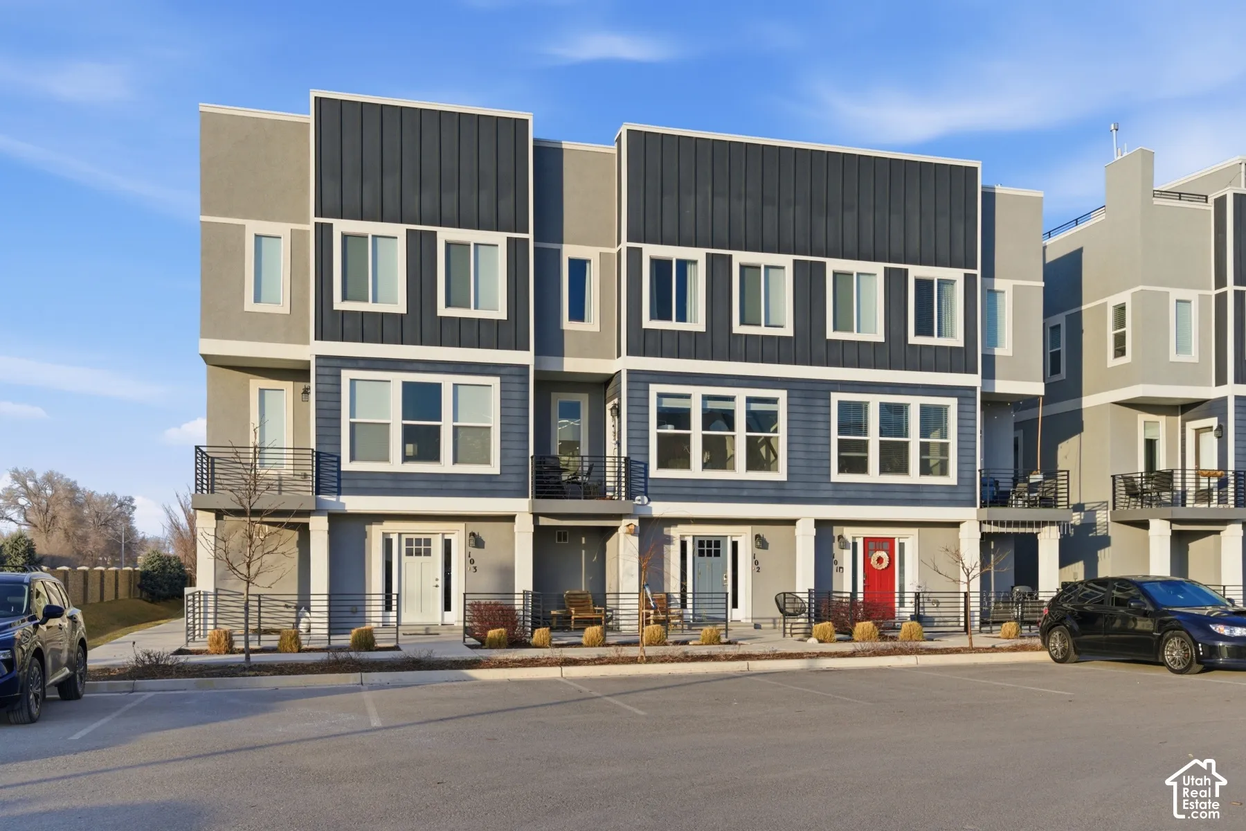 Contemporary townhome exterior featuring multi-textured siding, vertical metal panel accents, second-story balconies with horizontal cable railings, and private ground-level patios