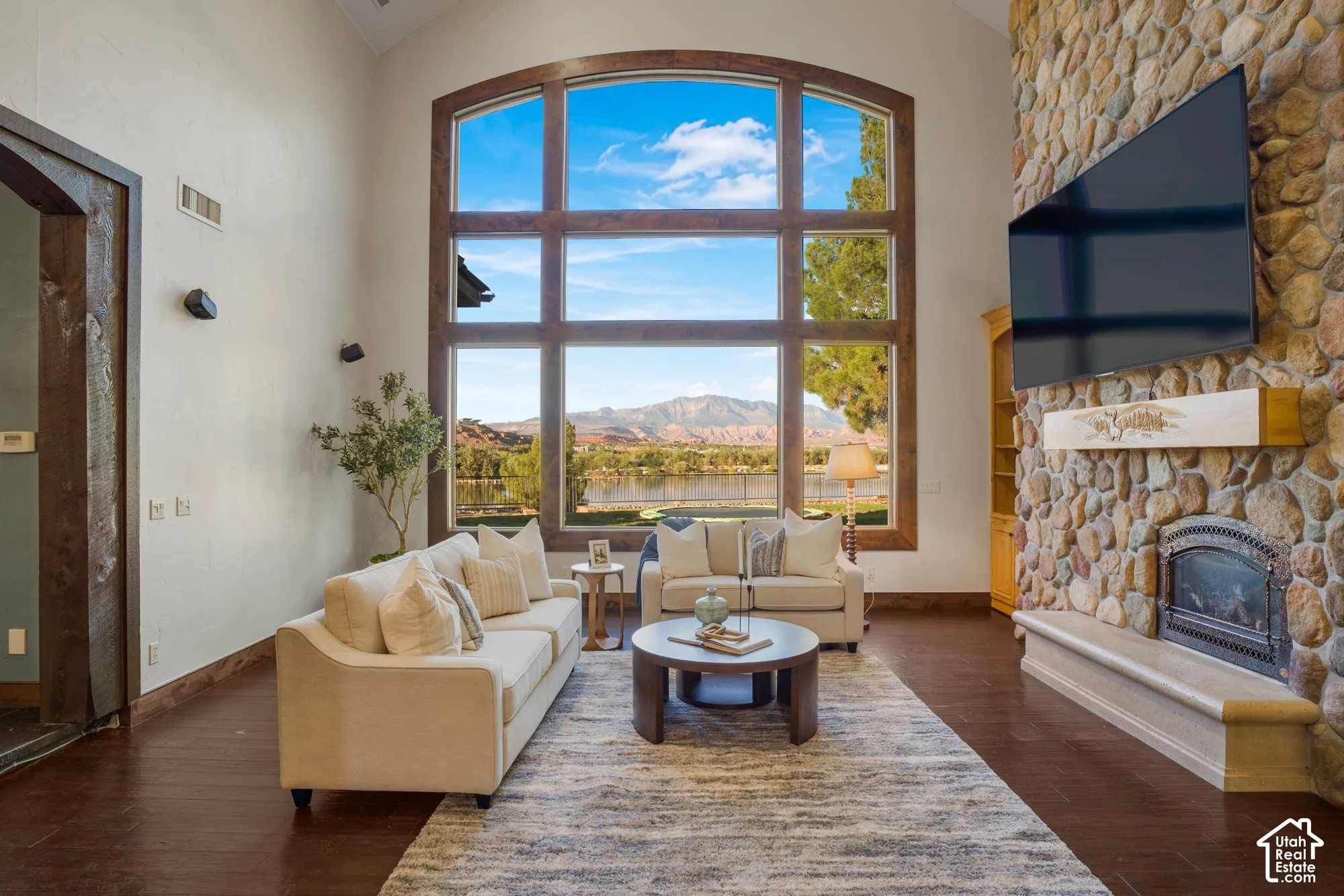 Vaulted ceilings, expansive multi-pane window with mountain and water scenery, natural stone fireplace surround, custom wooden mantel, and wood-finish flooring