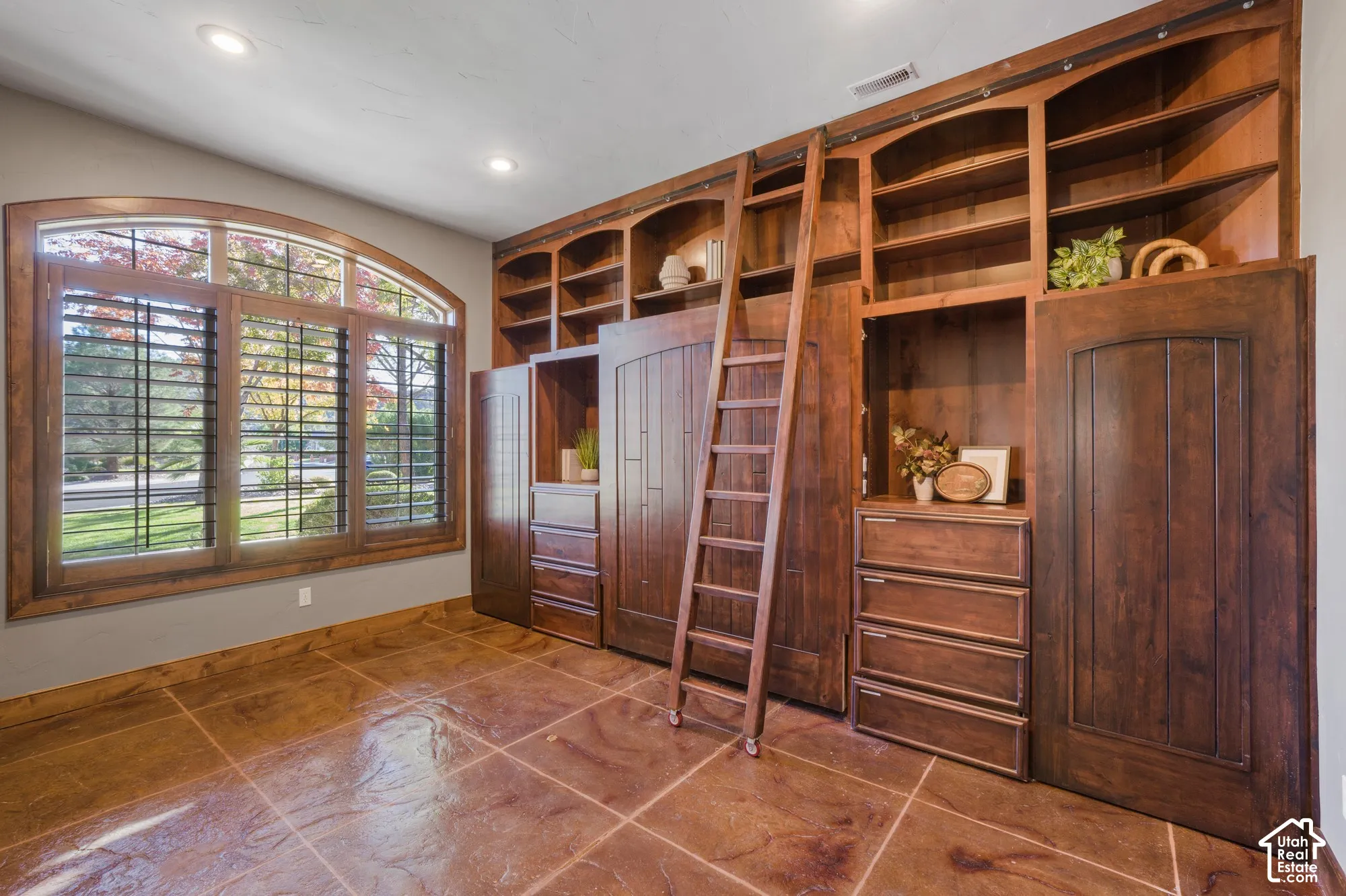 Custom floor-to-ceiling wooden cabinetry featuring a sliding ladder, integrated drawers, arched window with wood-frame trim, and stone-finish tile flooring