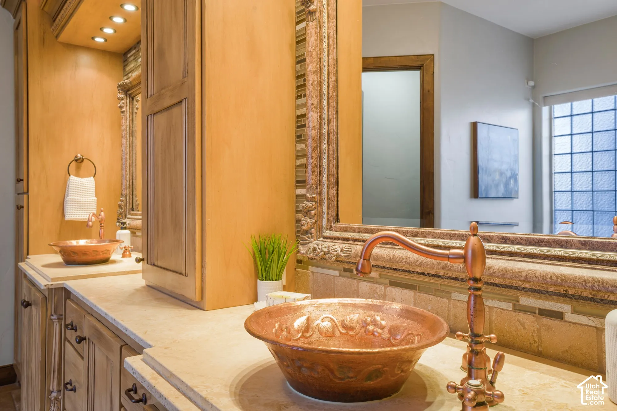 Custom wood cabinetry featuring integrated recessed lighting, stone countertop, dual hammered-copper vessel sinks with matching gooseneck faucets, and a large ornate framed mirror