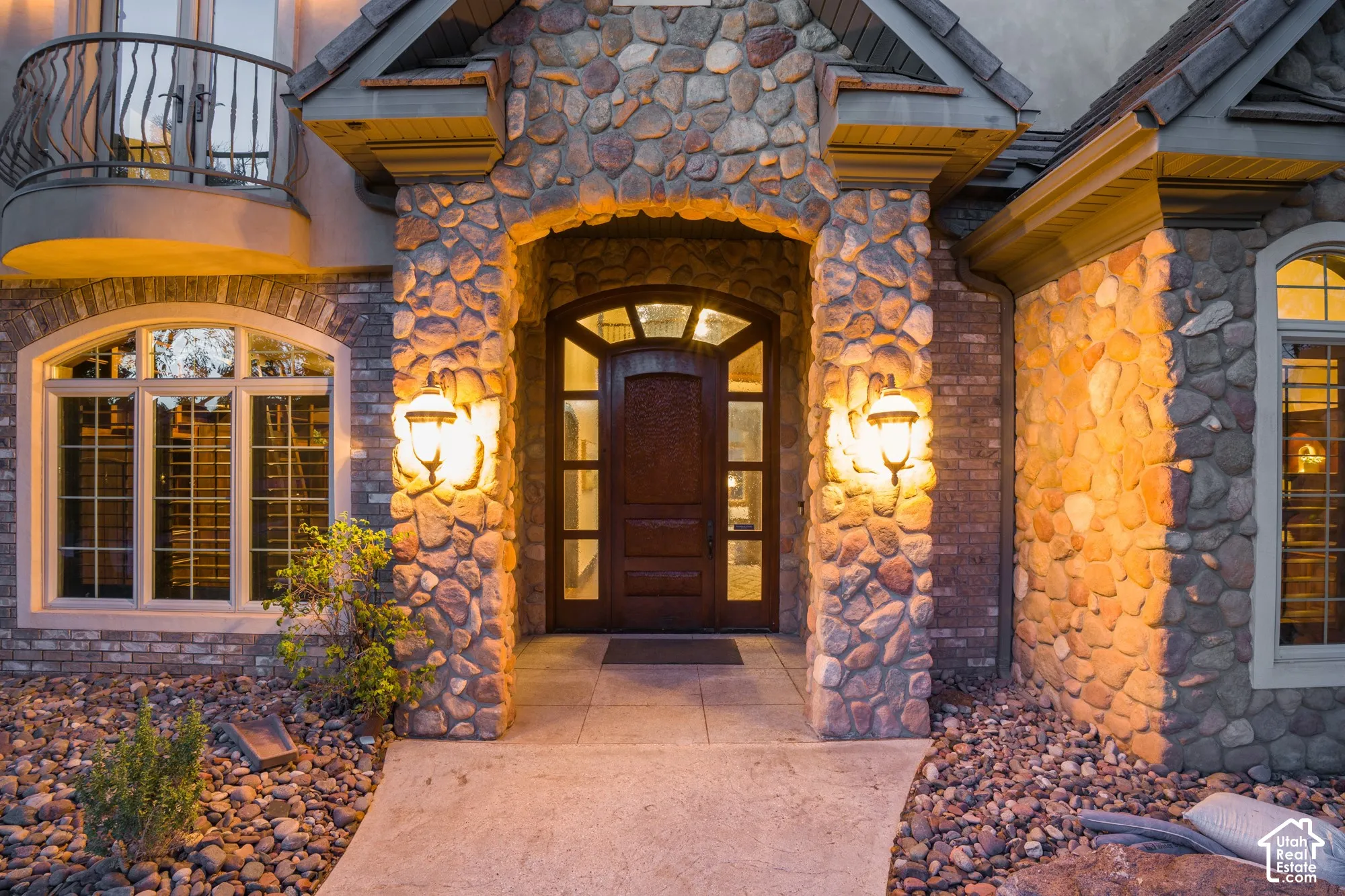 Stone veneer facade featuring a recessed entry, arched wood door with glass sidelights, decorative wall-mounted lighting, and a wrought iron balcony