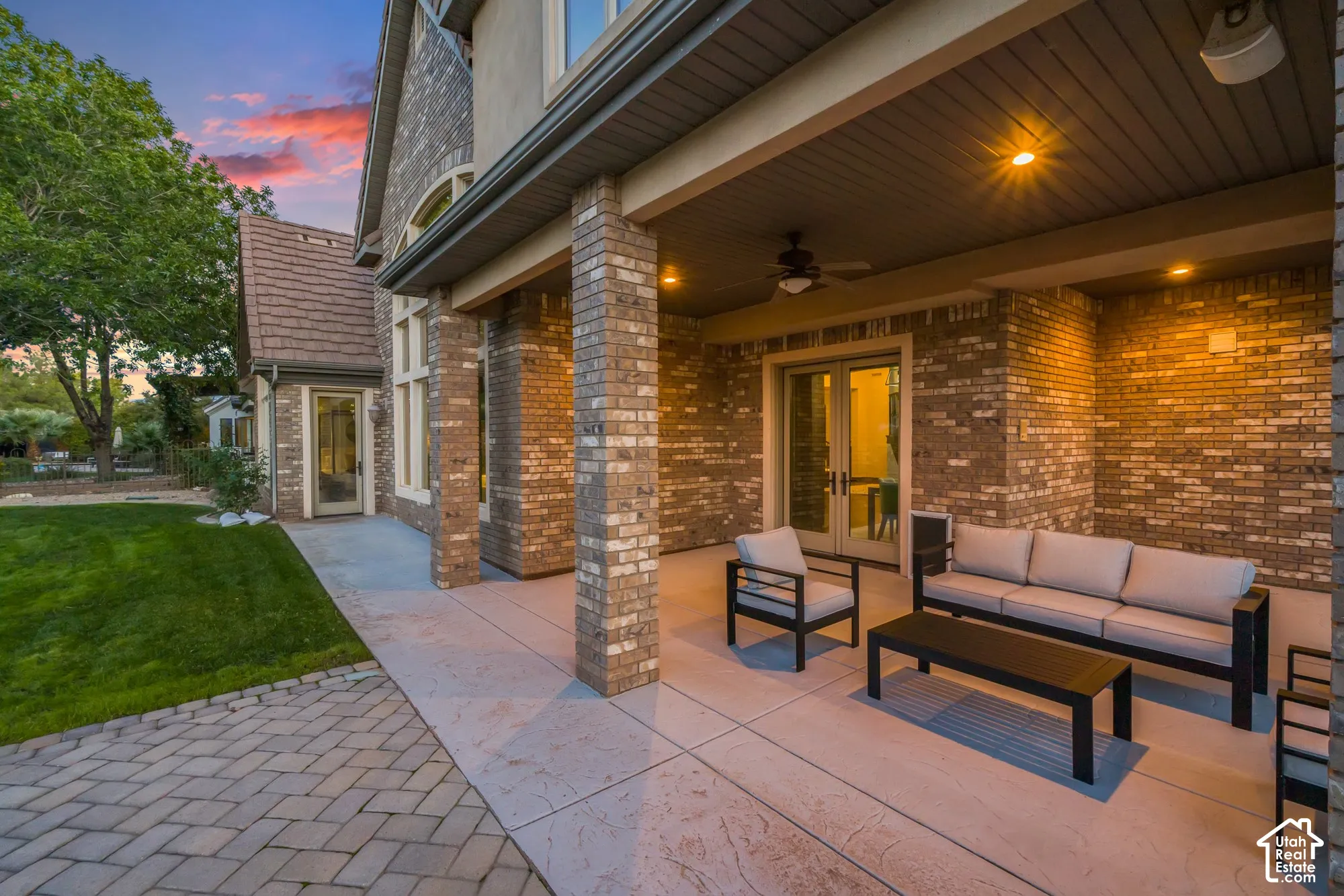 Covered brick patio featuring stamped concrete flooring, integrated ceiling fan, and recessed lighting, extending to a paver stone walkway and manicured lawn