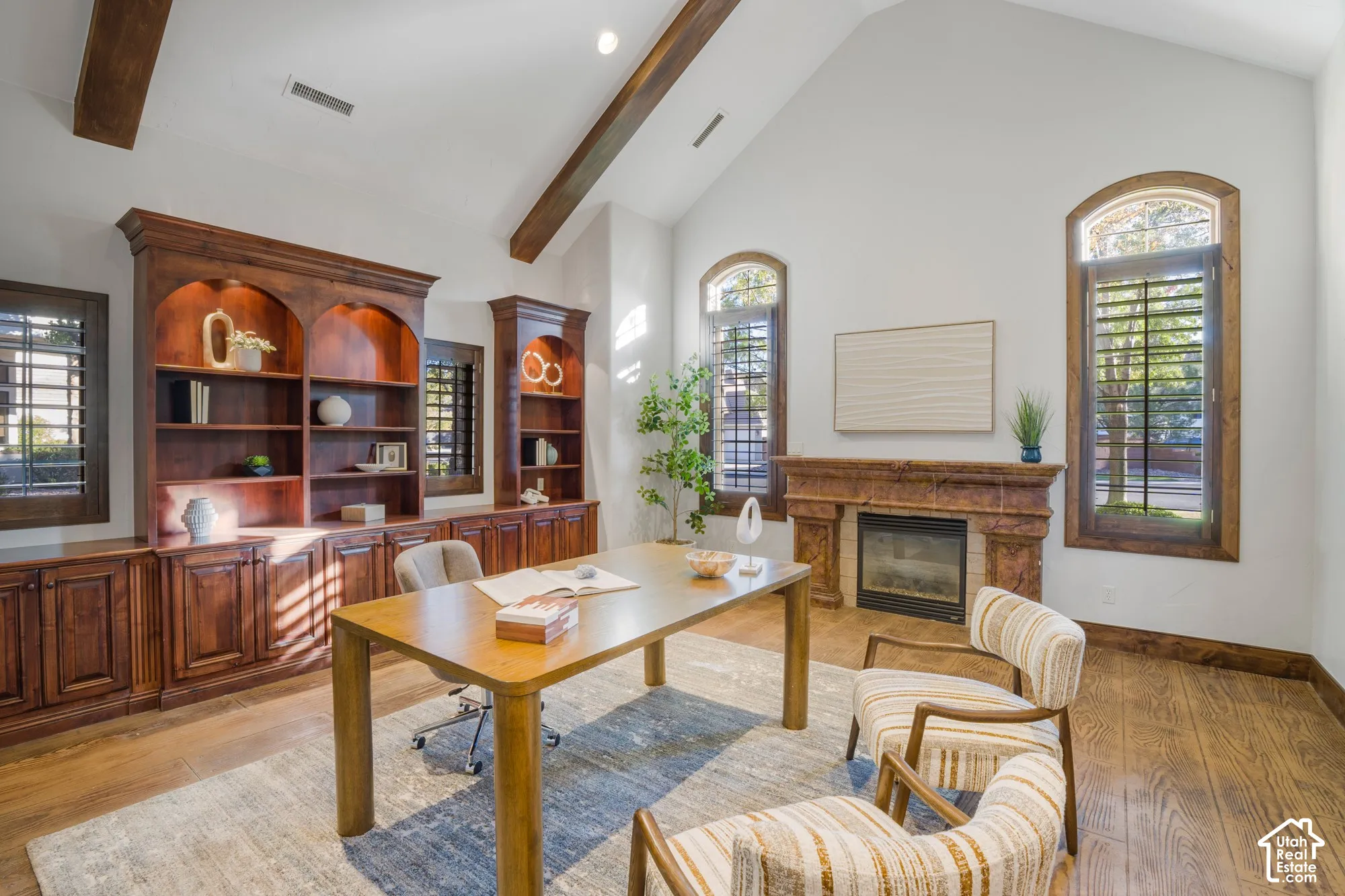 Vaulted ceiling with exposed wood beams, custom floor-to-ceiling wooden built-in cabinetry, arched windows with shutters, and a stone-surround fireplace
