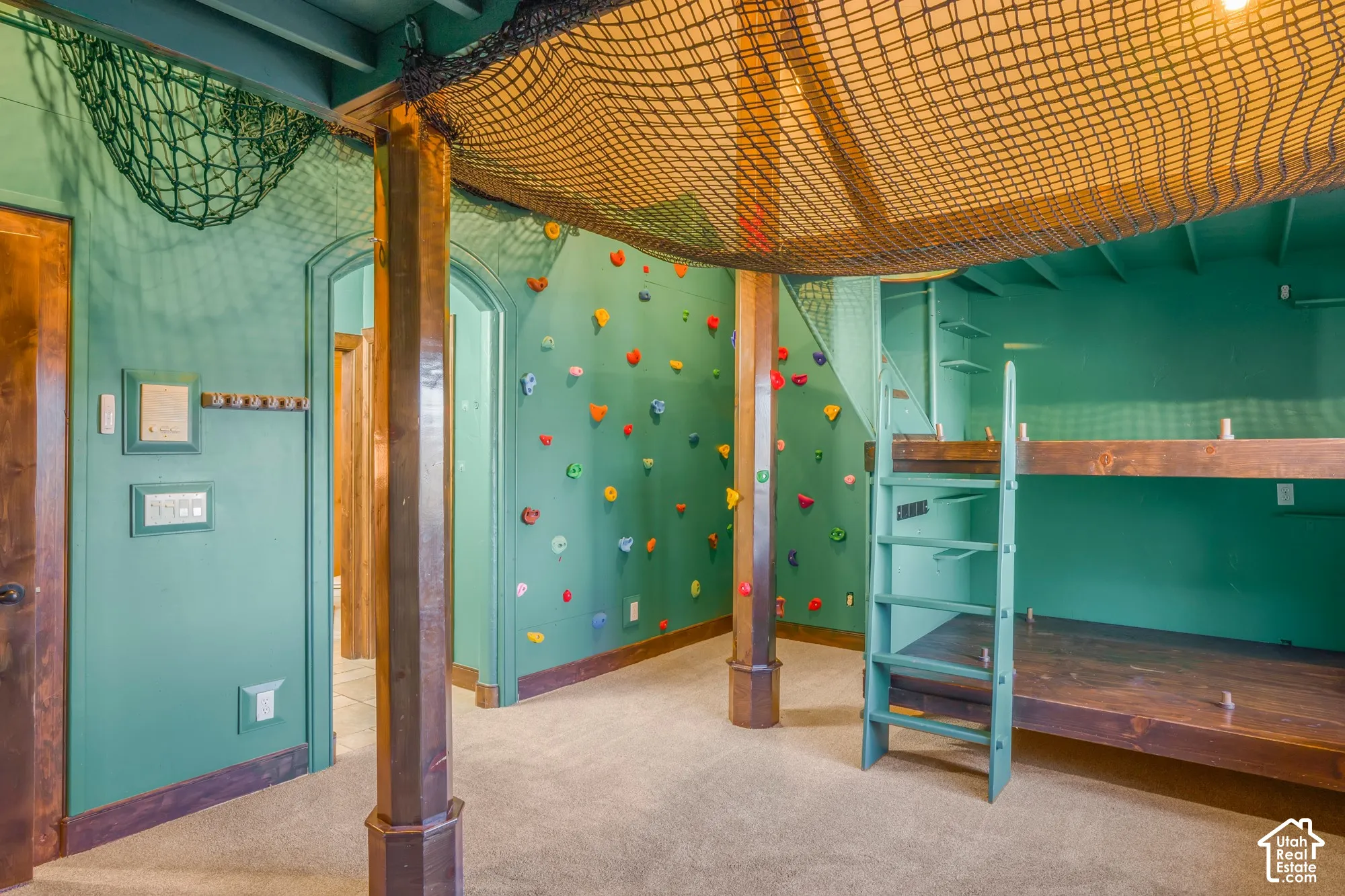 Built-in climbing wall with multi-colored holds, custom wood-framed bunk structure with integrated ladder, overhead cargo-style netting, and exposed wooden support columns