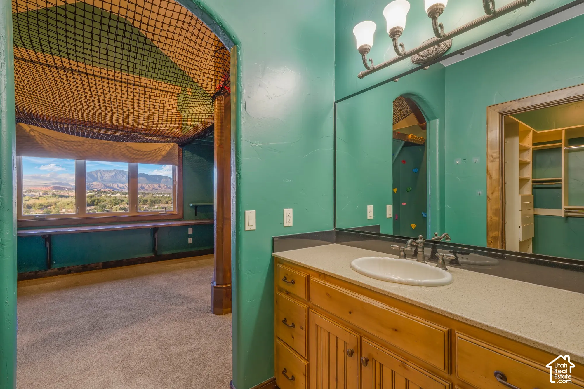 Bathroom vanity featuring wood cabinetry, a solid surface countertop with an integrated oval sink, and a large-format vanity mirror with attached light fixture, accompanied by an open space showcasing a mountain backdrop and built-in closet shelving