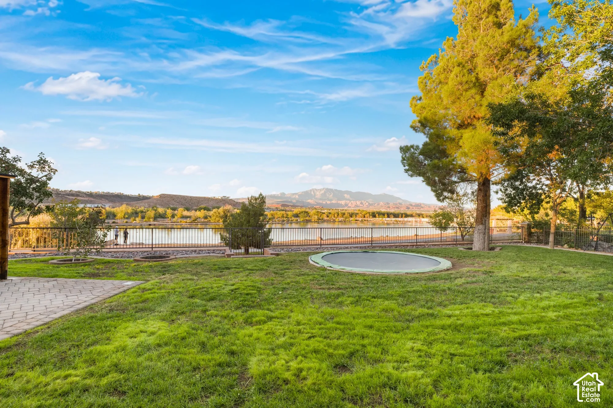 Expansive backyard featuring a manicured lawn, stone paver patio, mature trees, metal perimeter fencing, and mountain scenery