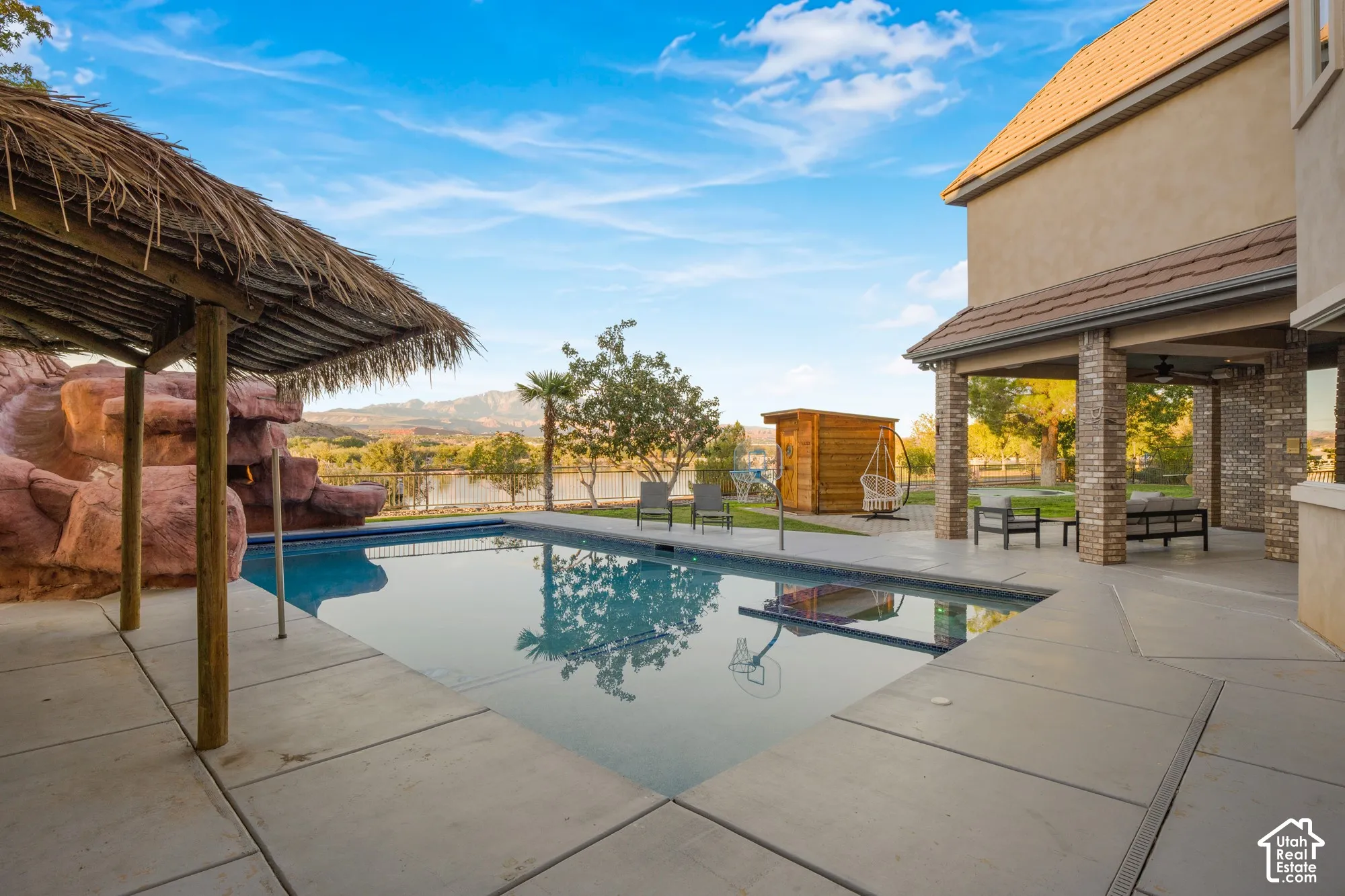 Backyard featuring a swimming pool, decorative rock water feature, thatch-roofed cabana, concrete patio, and covered porch with brick columns