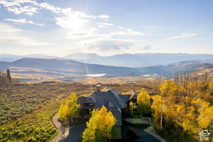 Multi-gabled residence with a shingled roof, stone chimney, paved driveway, and established foliage, set against an expansive mountain backdrop