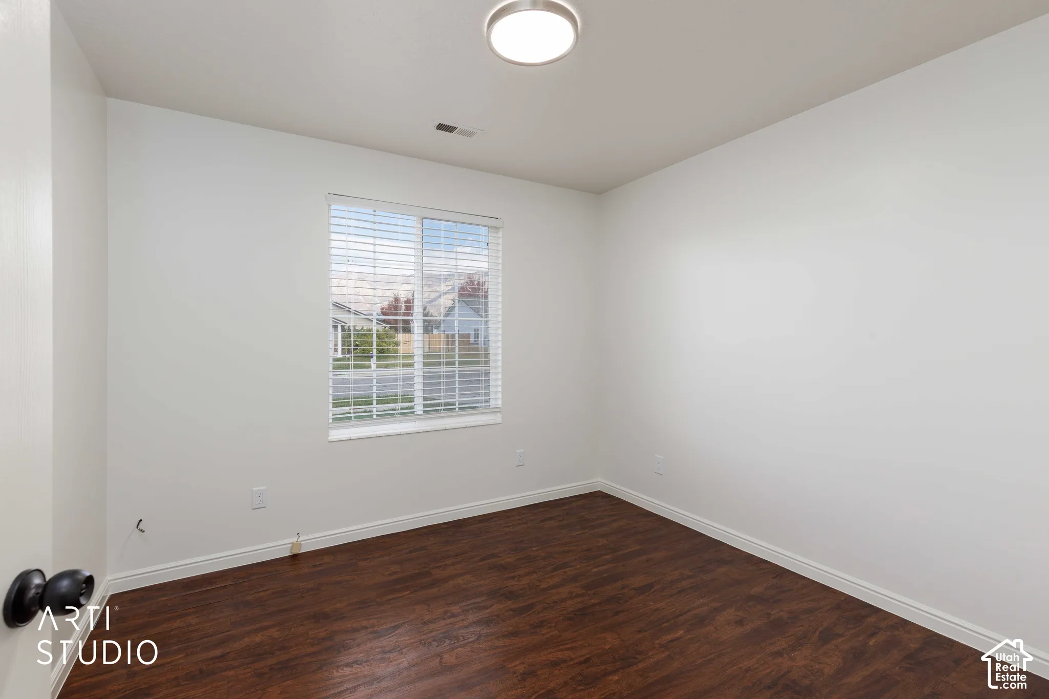 Interior room featuring dark wood-finish flooring, neutral white walls, a flush-mount circular ceiling light, and a window with horizontal blinds