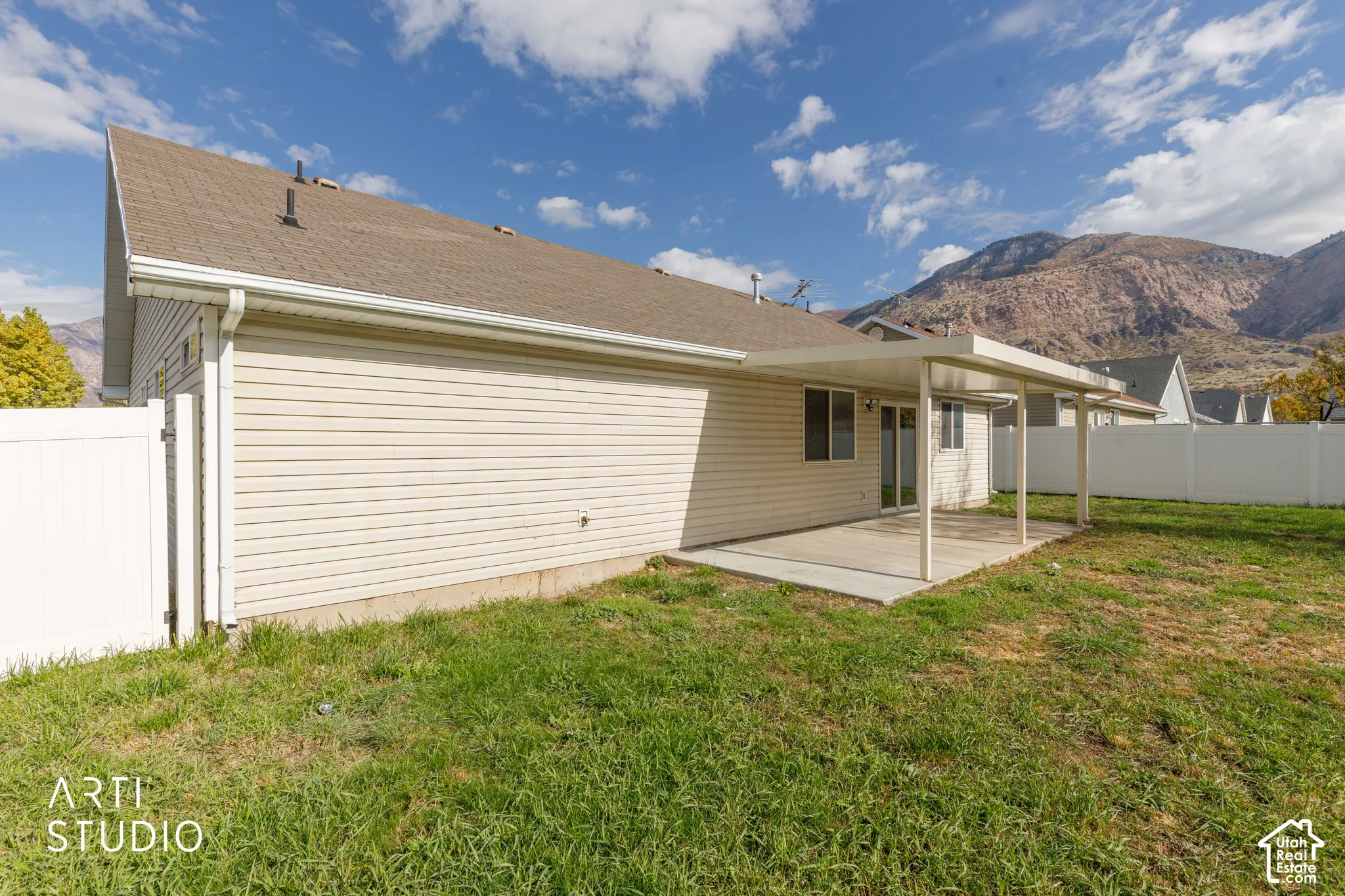 Rear exterior featuring horizontal vinyl siding, a concrete patio, a covered metal awning, a white privacy fence, and a mountain backdrop