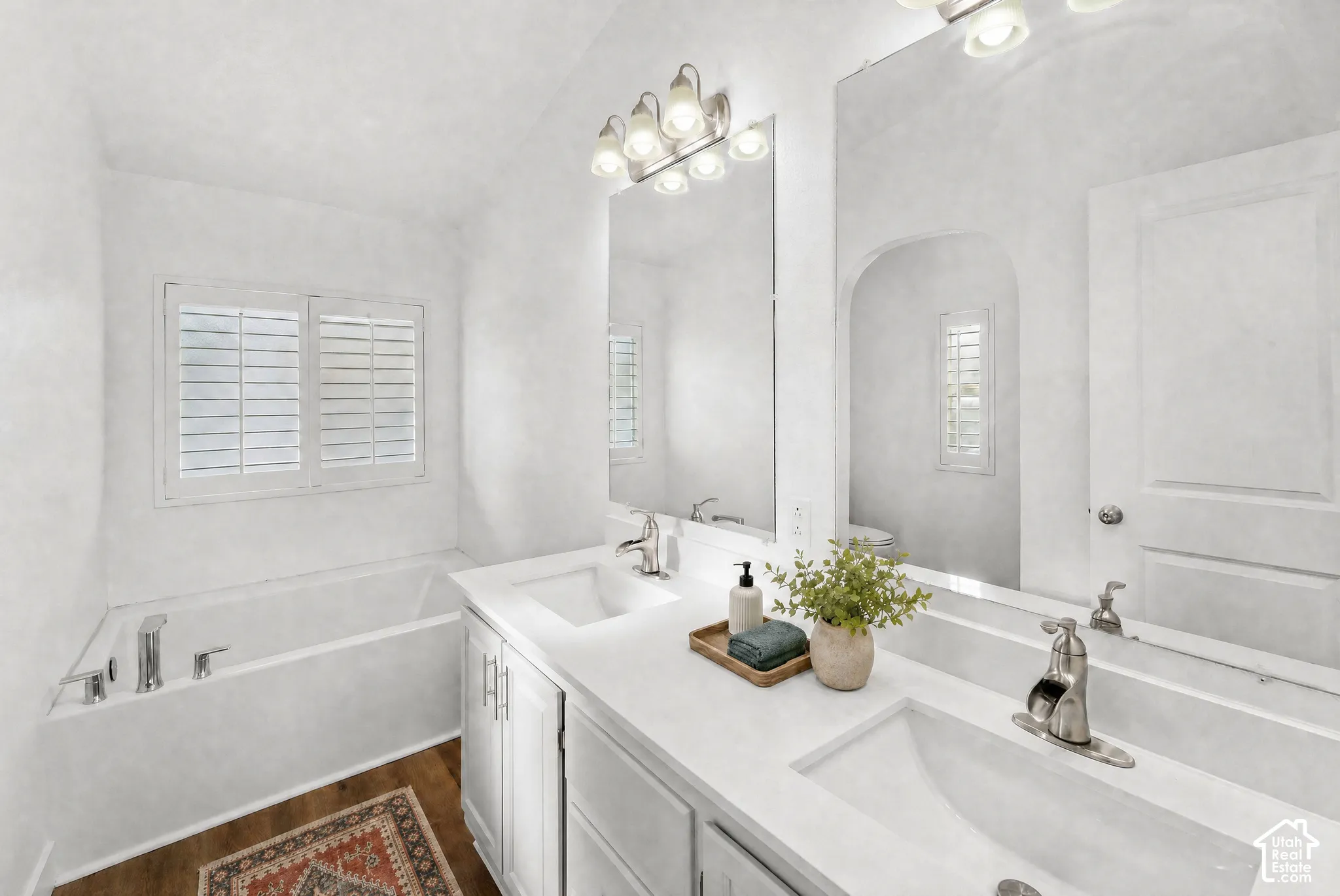 Bathroom featuring a dual-sink vanity with white cabinetry, a composite countertop, polished metal fixtures, and plantation shutters