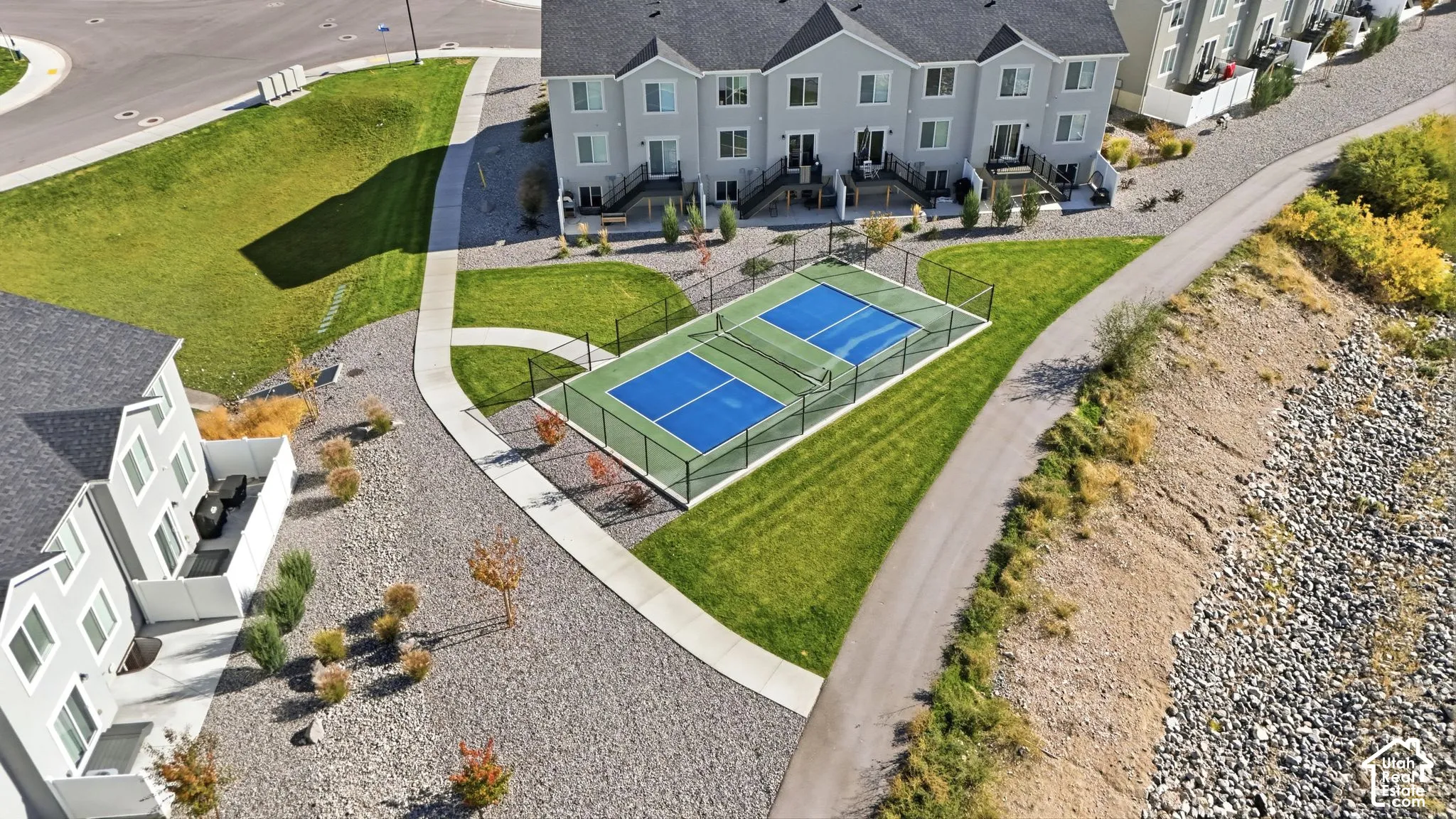 Multi-family residential complex featuring a paved pickleball court, fenced perimeter, concrete walkways, landscaped grounds, and paved pathways